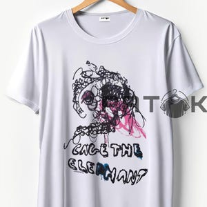 May include: White t-shirt with a black and pink abstract design and the text "CAGE THE ELEPHANT" in black. The shirt is hanging on a wooden hanger.