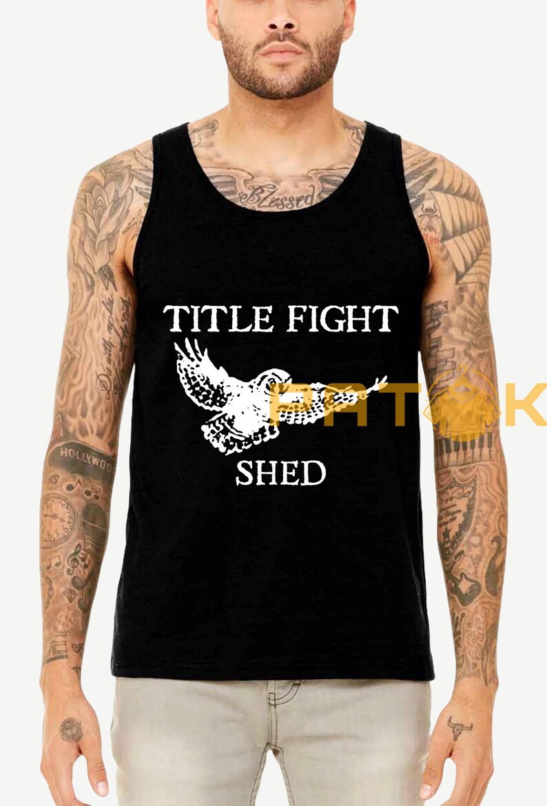 May include: Black vest top featuring the text "TITLE FIGHT SHED" in white, and a white graphic of an owl. The top is sleeveless with a rounded neckline. The owl graphic has outstretched wings.