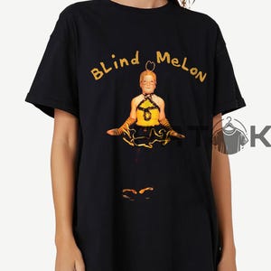 Puede incluir: Camiseta negra con la inscripción "BLIND MELON" en dorado sobre una imagen de una persona con un atuendo amarillo y negro. La persona está sentada con los brazos extendidos. Una camiseta gráfica con temática musical.