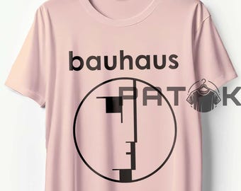 May include: A light pink t-shirt with the word "bauhaus" in black, above a black geometric design inside a circle. The shirt is hanging on a wooden hanger.