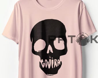 May include: A light pink t-shirt featuring a large black skull graphic. The skull has large eye sockets and the word "GOJIRA" is printed below the jawline. The shirt is hanging on a wooden hanger.