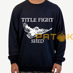 May include: Navy blue sweatshirt featuring a white owl graphic and the text "TITLE FIGHT SHED". The owl is shown with its wings spread. The sweatshirt is a long-sleeved crew neck.