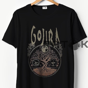 May include: Black t-shirt featuring the band name "GOJIRA" in a stylized font above a circular graphic. The graphic includes a tree of life design with a moon and other symbols. The shirt is hanging on a wooden hanger.