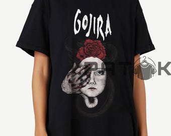 May include: Black t-shirt featuring the band name "GOJIRA" in white above a surreal graphic. The graphic includes a face with a red rose, a hand covering an eye, and a snake-like design.