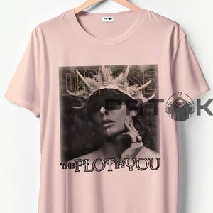 Puede incluir: Camiseta rosa claro con una imagen en blanco y negro de una persona con una corona. Las palabras "DISGUISE" y "THE PLOT IN YOU" están impresas en oro y negro.