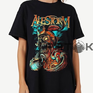 May include: Black t-shirt featuring the band name "ALESTORM" in orange and gold lettering. The graphic depicts a skull-faced figure with horns, feathers, and flowing hair in shades of orange, blue, and teal.