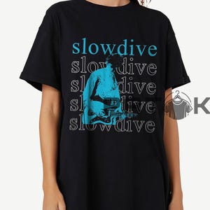 Puede incluir: Camiseta negra con el nombre de la banda "slowdive" en texto azul y blanco. Un gráfico azul y blanco de una persona tocando la guitarra está centrado en la camiseta. La camiseta es de estilo casual y unisex.