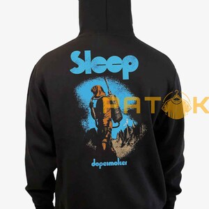 Pu&ograve; includere: Felpa con cappuccio nera con la scritta "Sleep" in blu a blocchi sopra una grafica di una persona in tuta arancione con uno zaino. La parola "dopesmoker" &egrave; sotto in blu.