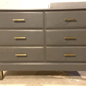 May include: A gray six-drawer dresser with gold-colored drawer pulls and legs. The dresser has a rectangular shape and a simple, modern design. The drawers are arranged in two columns of three.