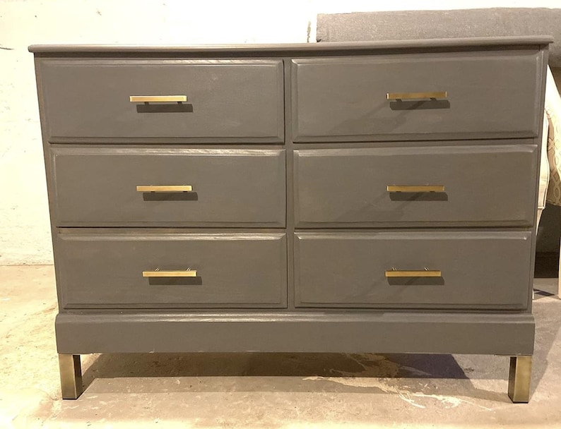 May include: A gray six-drawer dresser with gold-colored drawer pulls and legs. The dresser has a rectangular shape and a simple, modern design. The drawers are arranged in two columns of three.