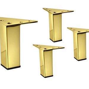 May include: Four gold-colored, triangular furniture legs. Each leg has a rectangular base with a black bottom and a triangular top with pre-drilled holes for mounting. The legs are made of a shiny, reflective metal.