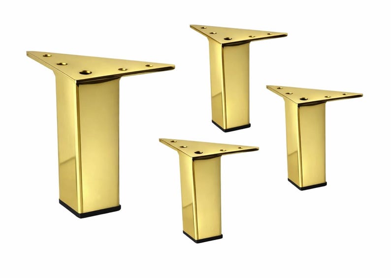 May include: Four gold-colored, triangular furniture legs. Each leg has a rectangular base with a black bottom and a triangular top with pre-drilled holes for mounting. The legs are made of a shiny, reflective metal.