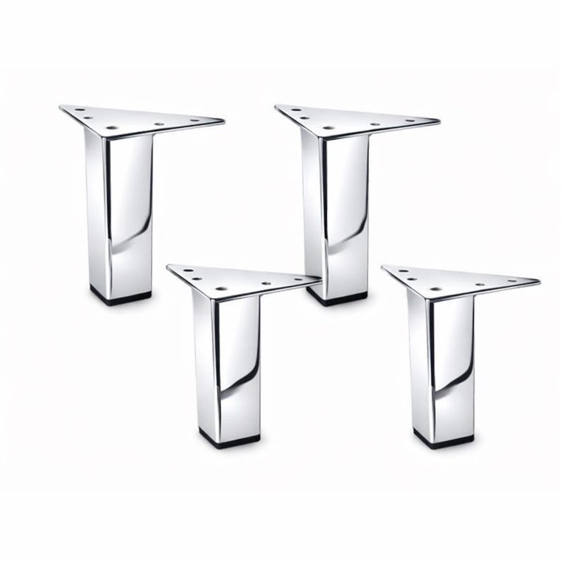May include: Four chrome-finished furniture legs. Each leg has a triangular top with screw holes and a rectangular base. The legs are reflective, with a shiny, polished appearance. The bases are black.