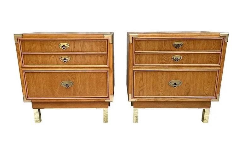 May include: Two matching wooden nightstands with three drawers each. The nightstands have brass hardware and corner accents. The legs are also brass. The wood has a warm, medium tone.