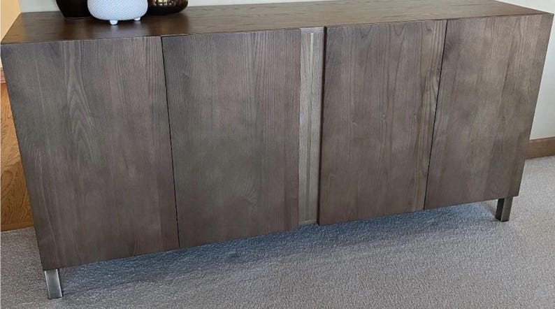May include: A modern, dark wood cabinet with four doors and brushed metal legs. The cabinet has a natural wood grain finish and a rectangular shape. Decorative items sit on top.