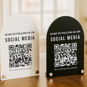 Könnte beinhalten: Zwei gewölbte Social-Media-Schilder, eines weiß und eines schwarz, mit dem Text "SCANNE UNS AUF SOCIAL MEDIA" und einem QR-Code. Die Schilder zeigen auch den Benutzernamen "@CraftSignShop2025".