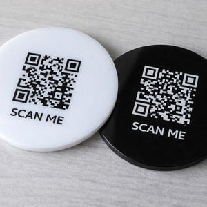 Pode incluir: Duas bases redondas, uma branca e outra preta, cada uma com um código QR e o texto "SCAN ME". As bases estão sobre uma superfície cinza claro.