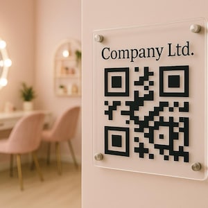 May include: A clear acrylic sign displaying a black QR code and the text "Company Ltd." is fixed to a pink wall. The sign is secured with silver fixings. The background features a vanity with a lit mirror and pink chairs.
