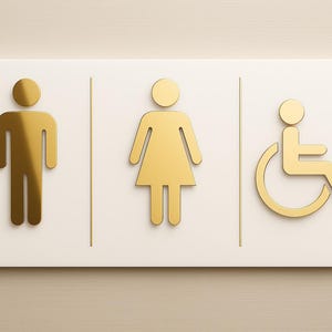 Puede incluir: Un letrero rectangular de baño con símbolos dorados de hombre, mujer y usuario de silla de ruedas. Los símbolos están sobre un fondo blanco, separados por líneas verticales. El letrero está montado en una pared beige claro.