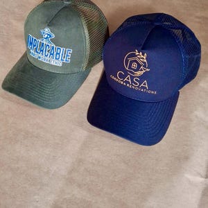 May include: Two trucker hats. One is olive green with a mesh back and the text "IMPLACABLE HARD WORK ETHIC". The other is navy blue with a mesh back and the text "CASA CORDOBA RENOVATIONS".
