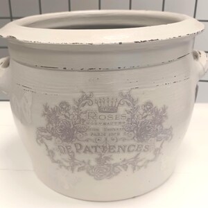 Peut inclure: Un pot de fleurs en céramique blanc cassé vieilli, avec un large bord et deux petites poignées latérales. Il présente un motif floral estompé avec les mots "Roses" et "De Patience". Le pot a un aspect rustique et vintage.