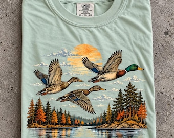 May include: A light green crew neck t-shirt featuring a graphic of three ducks flying over a lake and forest at sunset. The shirt has a Comfort Colors brand label at the neck with size XL printed on it. The graphic includes orange and green tones.