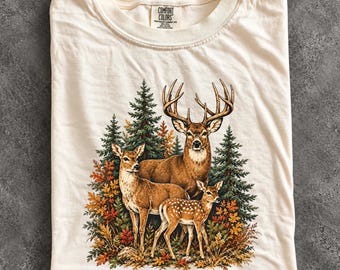 May include: An ivory crew neck t-shirt featuring a graphic print of a buck, a doe, and a fawn standing in a forest of pine trees and autumn foliage. The inner neck label displays the text Comfort Colors, Heavyweight Garment Dye, 100% Cotton.