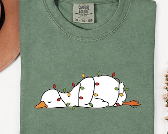 May include: A sage green crew neck t-shirt featuring a graphic of a white duck lying down, wrapped in colourful Christmas string lights. The shirt has a Comfort Colors brand label at the inner neckline with the size XL-TG-EG clearly printed.