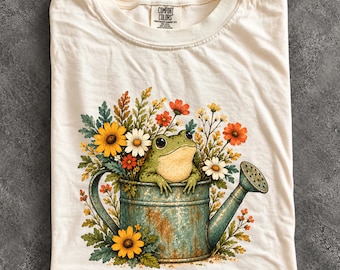 May include: An ivory t-shirt featuring a graphic of a green frog sitting inside a weathered teal watering can surrounded by colourful wildflowers including yellow daisies, orange blossoms, and white flowers. The neck label reads Comfort Colors.