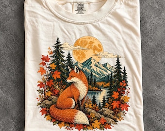 May include: An ivory t-shirt featuring a graphic print of a fox sitting in a forest with autumn leaves, pine trees, a lake, mountains, and a large full moon in the background. The inner neck label displays the text Comfort Colors.