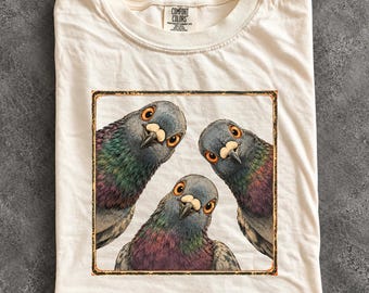 May include: An ivory t-shirt featuring a square graphic print of three pigeons looking directly forward. The pigeons have iridescent purple and green neck feathers, orange eyes, and grey beaks. The graphic is framed by a thin, distressed border.