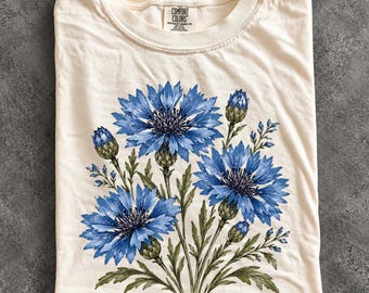May include: An ivory short-sleeve t-shirt featuring a graphic print of blue cornflowers with green stems and leaves. The inner neck label displays the text Comfort Colors, Heavyweight Garment Dye, 100% Cotton. The word Ivory is printed in the bottom left corner.