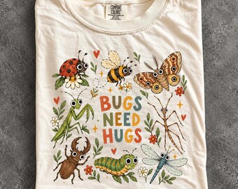 May include: An ivory t-shirt featuring a colourful graphic of various insects including a ladybug, bee, moth, praying mantis, stick insect, beetle, caterpillar, and dragonfly. The text "BUGS NEED HUGS" is printed in the centre in multi-coloured letters.