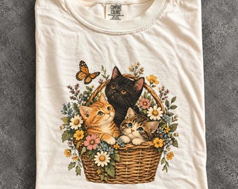 May include: An ivory t-shirt featuring a graphic print of three kittens—one orange tabby, one black, and one brown tabby—sitting inside a woven wicker basket filled with colourful wildflowers and a single monarch butterfly.
