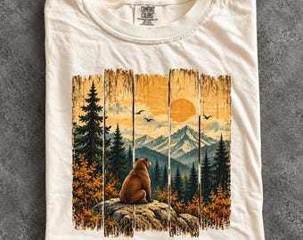 May include: An ivory t-shirt featuring a graphic print of a brown bear sitting on a rock, overlooking a mountain range, pine forest, and a sunset sky. The design is segmented by vertical lines and includes the Comfort Colors brand label.