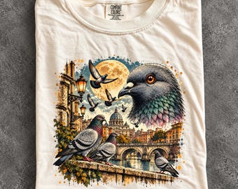 May include: An ivory t-shirt featuring a graphic print of pigeons in a city setting. The design includes a large pigeon head, several flying and perched pigeons, a full moon, street lamps, and a domed building. The label reads Comfort Colors.
