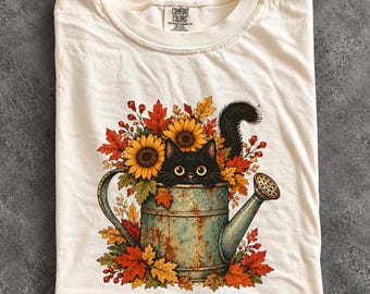 May include: An ivory t-shirt featuring a graphic of a black cat peeking out from a rustic, weathered blue-green metal watering can. The cat is surrounded by vibrant autumn foliage, including orange and yellow maple leaves, sunflowers, and red berries.