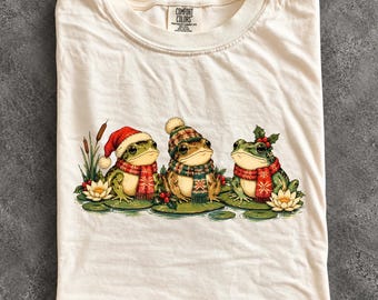 May include: An ivory t-shirt featuring a graphic of three green frogs sitting on lily pads. The frogs wear winter accessories including a red Santa hat, a patterned knit beanie, and a holly sprig. Each frog wears a red and green patterned scarf.