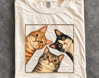 May include: An ivory t-shirt featuring a square graphic of three cats looking upward. The cats include an orange tabby, a black and white cat, and a brown tabby. The shirt has a Comfort Colors brand label at the inner neckline.