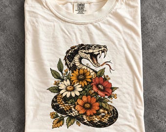 May include: An ivory t-shirt featuring a graphic print of a coiled snake with its mouth open, surrounded by a cluster of orange, red, and white flowers with green leaves. The neck label reads Comfort Colors, Heavyweight Garment Dye.