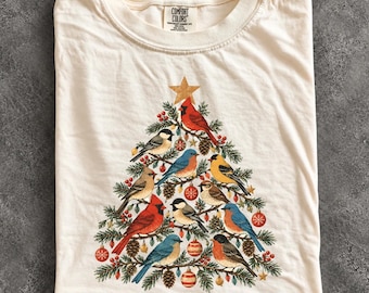 May include: An ivory crew neck t-shirt featuring a graphic of a Christmas tree formed by various birds, including cardinals and bluebirds, decorated with red and gold ornaments, pinecones, and a gold star at the top. The inner label reads Comfort Colors.