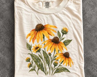 May include: An ivory t-shirt featuring a graphic print of yellow coneflowers with dark brown centres and green leaves. The inner neck label displays the text Comfort Colors, Heavyweight Garment Dye, 100% Cotton.