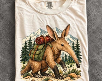 May include: An ivory t-shirt featuring a graphic of an aardvark wearing a green backpack with a red plaid blanket roll. The aardvark is standing in a landscape with pine trees and snow-capped mountains. The shirt has a Comfort Colors label.