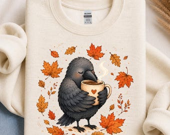 May include: A cream-coloured crewneck sweatshirt featuring a graphic of a black crow holding a beige mug with a small red heart. The crow is surrounded by scattered orange and red autumn leaves. The Gildan brand tag is visible at the inner neckline.