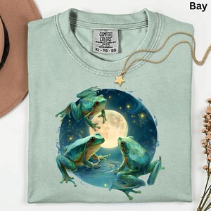 May include: A light green Comfort Colors XL-TG-EG sweatshirt featuring a graphic of three turquoise frogs around a full moon. A gold star pendant necklace rests on the shirt. The shirt is folded.