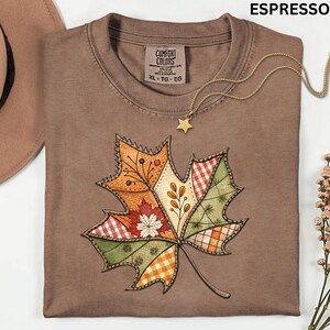 May include: Espresso-colored t-shirt with a large, colorful leaf design. The leaf is composed of various patchwork patterns in orange, green, and red. A gold star pendant necklace rests on the shirt. The shirt is labeled XL-TG-EG.