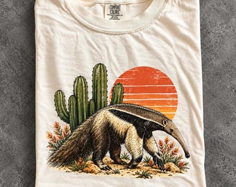 May include: An ivory t-shirt featuring a graphic of an anteater walking in a desert landscape with green cacti, small orange flowers, and a large orange sun. The inner neck label displays the text Comfort Colors, Heavyweight Garment Dye, 100% Cotton.
