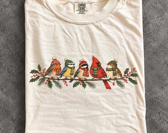 May include: An ivory t-shirt featuring a graphic of five birds perched on a holly branch. Each bird wears a knit hat and scarf. The birds include a robin, blue tit, chickadee, cardinal, and sparrow. The Comfort Colors brand label is at the neck.