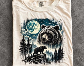 May include: An ivory t-shirt featuring a graphic print of a large bear head and a smaller bear standing on a rocky cliff. The scene includes a full moon, a starry night sky, and a dense pine forest. The shirt has a Comfort Colors brand label.