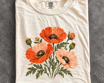 May include: An ivory short-sleeve t-shirt featuring a graphic print of orange and peach-coloured poppy flowers with green stems and leaves. The inner neck label displays the text Comfort Colors, Heavyweight Garment Dye, 100% Cotton.